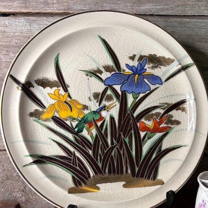 May include: Cream-colored ceramic plate with a gold rim and a floral design. The plate features yellow, blue, and orange iris flowers, with a small bird perched among the dark brown and green leaves. The background has gold accents.