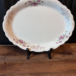 May include: Oval, white serving platter with a scalloped edge. The platter features floral designs in shades of purple and green, with accents of gold. The platter is displayed on a black stand, set against a wooden background.