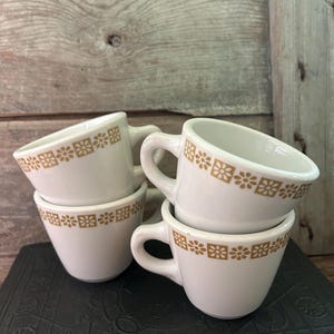 May include: Three white ceramic mugs featuring a repeating gold-colored floral pattern around the upper rim. Each mug has a curved handle and a tapered base. The mugs are stacked and arranged together, showcasing the consistent design on each piece.