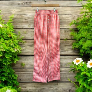 May include: Red and white gingham-patterned pants hanging on a wooden hanger against a wooden fence background. The pants have a button-up closure and a high waist. Green foliage and white flowers frame the pants.