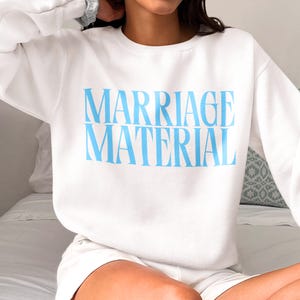 May include: White sweatshirt with blue text that reads "MARRIAGE MATERIAL".