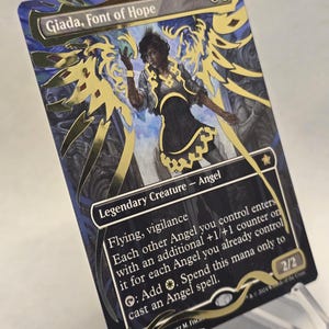 Pode incluir: Um cartão Magic: The Gathering com "Giada, Font of Hope". O cartão tem um fundo preto com detalhes dourados e uma ilustração de um anjo. O texto inclui "Legendary Creature - Angel" e instruções do jogo. O cartão é exibido em um suporte transparente.