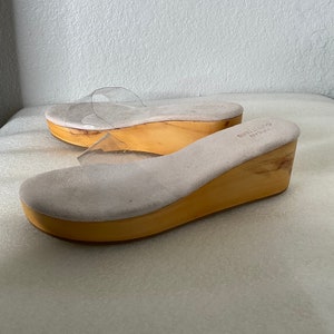May include: A pair of clear plastic heeled sandals with a wooden platform. The sandals have a light brown suede insole and a light brown wooden platform.