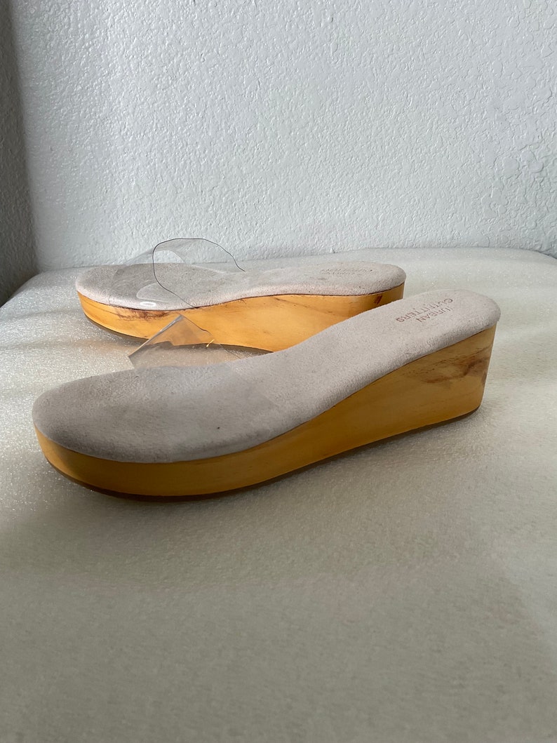 May include: A pair of clear plastic heeled sandals with a wooden platform. The sandals have a light brown suede insole and a light brown wooden platform.
