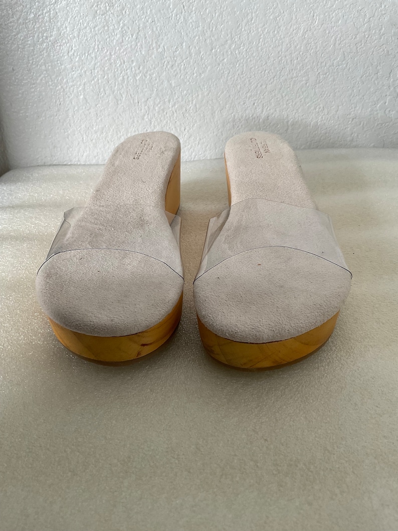 May include: A pair of white platform sandals with clear plastic straps. The sandals have a wooden platform and a suede upper.