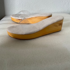 May include: A pair of beige suede wedge sandals with clear plastic straps. The sandals have a wooden wedge heel.