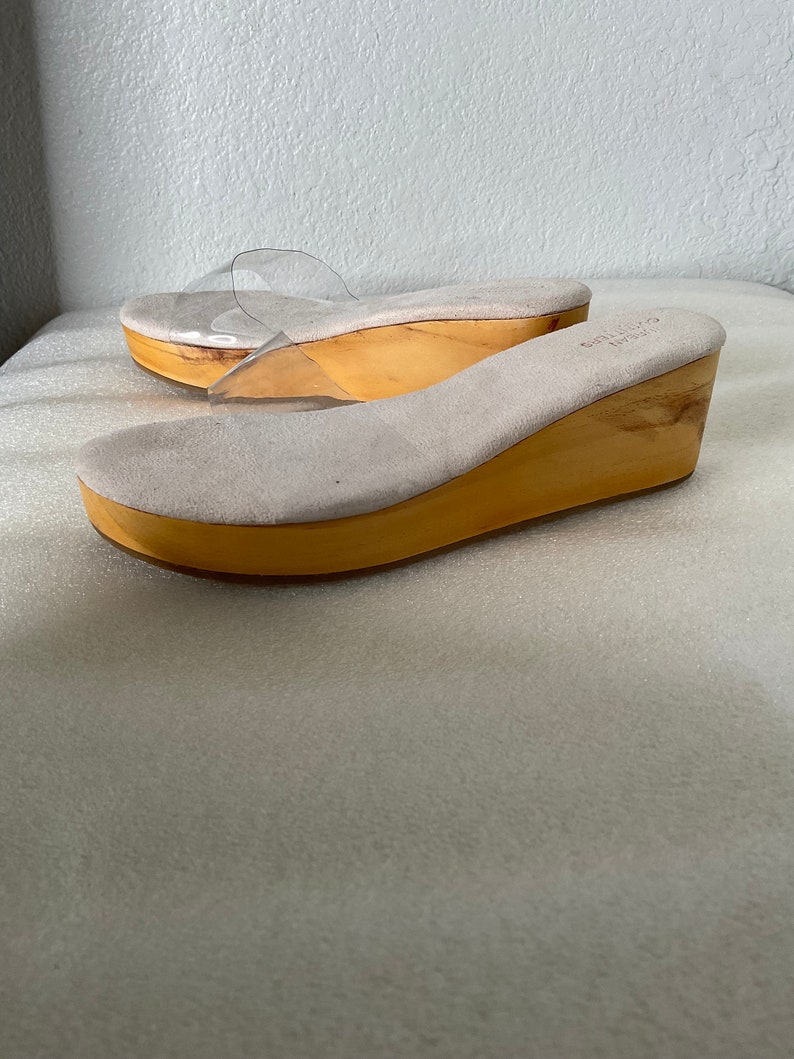 May include: A pair of beige suede wedge sandals with clear plastic straps. The sandals have a wooden wedge heel.