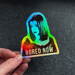 Könnte beinhalten: Holografischer Aufkleber mit Regenbogenverlauf, der eine Frau und den Text "BORED NOW" zeigt. Der Aufkleber wird von einer Hand gehalten. Der Hintergrund ist dunkel.