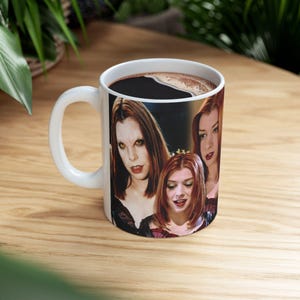 May include: White ceramic coffee mug featuring a group of red-haired women with dark makeup. The mug is filled with dark coffee and has a white handle. The mug is sitting on a wooden surface.