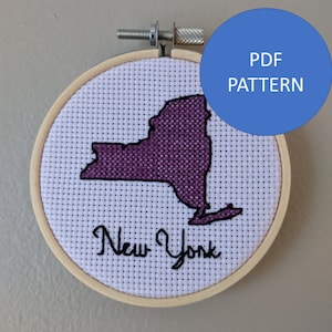 May include: A cross-stitch embroidery hoop with a purple outline of the state of New York and the text "New York" in black thread.