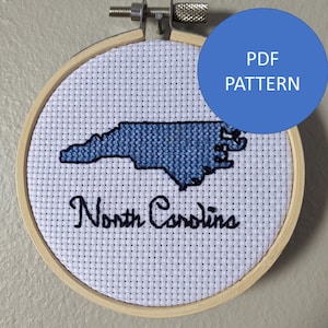 May include: A cross-stitch embroidery of the state of North Carolina in blue thread on white Aida cloth, mounted in a wooden embroidery hoop. The words "North Carolina" are stitched in black thread below the state outline. A blue circle with the text "PDF PATTERN" is in the top right corner of the image.