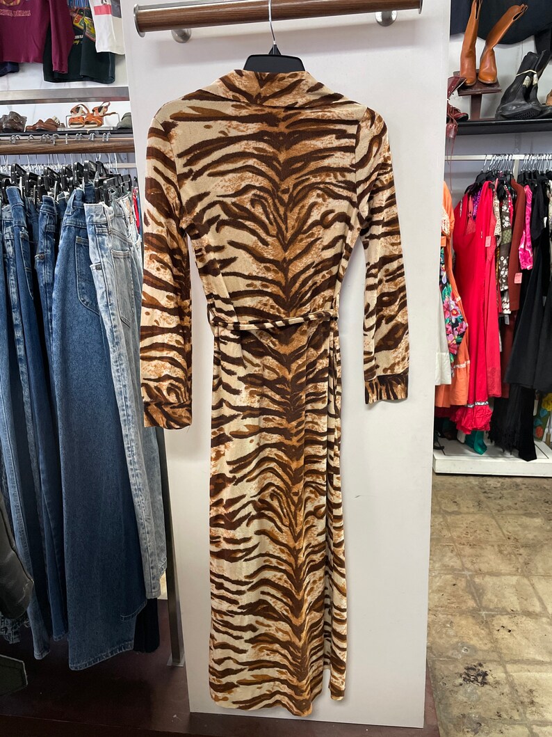 May include: A long, brown and white tiger print maxi dress with long sleeves and a tie belt. The dress is hanging on a rack in a clothing store.