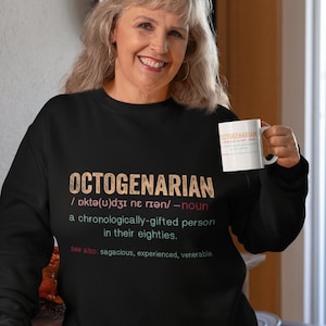 May include: A woman wearing a black sweatshirt with the word "Octogenarian" defined as a "chronologically-gifted person in their eighties." The sweatshirt has a vintage, distressed look.