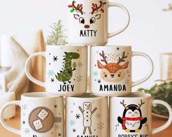 May include: Six white ceramic mugs stacked in a pyramid, each featuring a unique Christmas-themed illustration and a name. Designs include a reindeer, dinosaur, deer, hot cocoa, snowman, and penguin. Text reads: Matty, Joey, Amanda, Rowan, Samuel, Bobby's Mug.