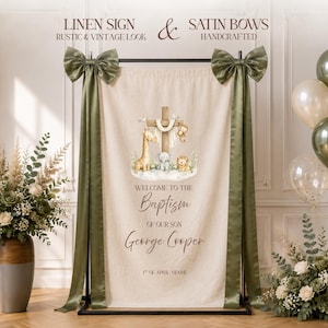 Pode incluir: Uma placa de linho bege pendurada num suporte de metal preto com laços de cetim verde-azeitona. A placa apresenta uma ilustração de uma cruz, girafa, elefante e leão com o texto: WELCOME TO THE Baptism OF OUR SON George Cooper 1ST OF APRIL MIAMI.