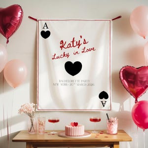 May include: A festive scene with pink balloons and heart-shaped foil balloons. A banner reads "Katy's Lucky in Love" with playing card symbols. A table displays a cake, drinks, and flowers, suggesting a bachelorette party.