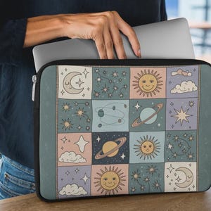 May include: A rectangular laptop sleeve featuring a grid pattern of celestial illustrations including smiling suns, crescent moons, planets with rings, stars, and clouds in muted shades of blue, pink, and cream with a black zipper border.