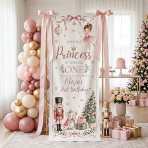 Op de afbeelding: Een verticale banner in roze en goud met een ballerina, een notenkraker, een muis en een kerstboom. De tekst luidt: Our Little Princess is Turning One, Welcome to Celebrate Olivia's First Birthday. Roze linten hangen aan de bovenhoeken en links staat een ballonnenboog.