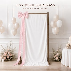 Puede incluir: Un gran lazo de satén rosa sujeto a un marco de madera cubierto con un telón de fondo de tela blanca. El texto dice HANDMADE SATIN BOWS AVAILABLE IN 24 COLORS. La escena incluye racimos de globos blancos y arreglos florales en el suelo.