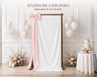 May include: A large pink satin bow attached to a wooden frame draped with a white fabric backdrop. The text reads HANDMADE SATIN BOWS AVAILABLE IN 24 COLORS. The scene includes clusters of white balloons and floral arrangements on the floor.