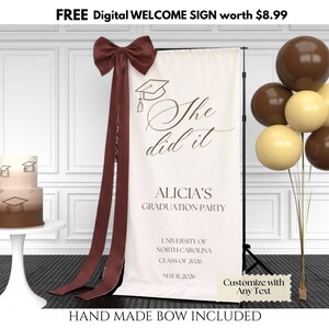 May include: A cream-coloured graduation banner with the text "She did it" and "Alicia's Graduation Party". It features a brown bow and ribbons. A two-tiered cake and balloons in brown and cream are also visible.