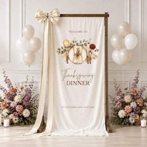May include: A vertical cream-colored fabric banner on a wooden stand, featuring a printed illustration of a place setting with a candle and berries. The text reads "WELCOME TO Thanksgiving DINNER 14TH NOVEMBER 2025, NEW YORK" with a large cream bow.