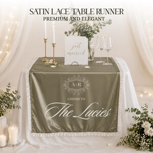 May include: A satin lace table runner in olive green with white lace trim, set for a wedding. The runner is on a white tablecloth, with text that reads "Cheers to The Lucies". The table is decorated with flowers, candles, and wine glasses.