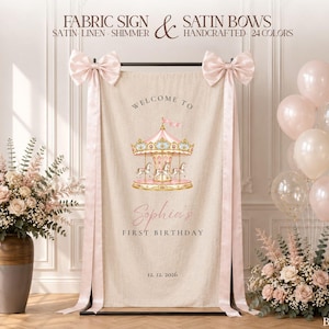 Peut inclure: Une enseigne rectangulaire en tissu beige suspendue sur un support noir, avec une illustration de carrousel, le texte "WELCOME TO Sophia's FIRST BIRTHDAY 12. 12. 2026" et deux grands nœuds en satin rose pâle aux coins supérieurs.