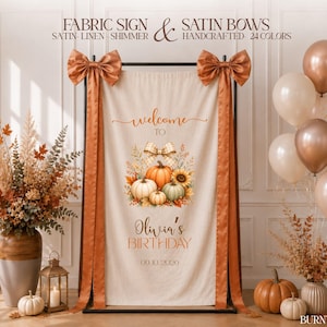 Puede incluir: Un letrero de tela color crema con un gráfico de calabazas y girasoles y el texto "welcome TO Olivia's BIRTHDAY 09.10.2026". El letrero cuelga de un marco de metal negro decorado con grandes lazos de satén naranja quemado y cintas largas.