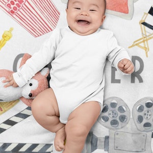May include: A smiling baby wearing a white onesie lies on a white blanket decorated with movie-themed illustrations. The blanket features popcorn, 3D glasses, a film clapperboard, a film reel, and an Oscar statue.
