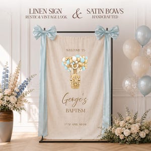 Pode incluir: Uma placa retangular de linho bege com um gráfico de balão de ar quente, um ursinho de pelúcia, balões dourados e azuis e o texto "WELCOME TO George's BAPTISM 1ST OF APRIL MIAMI". A placa é decorada com dois laços de cetim azul claro.