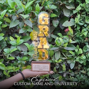 Puede incluir: Un trofeo de graduación de acrílico dorado con la palabra "GRAD" en letras grandes. El texto "Class of 2026" está en cursiva. El trofeo incluye una base de madera con detalles grabados: "Caroline Cooper, University of California, MBBS".