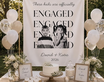 May include: A white fabric backdrop featuring the text These kids are officially ENGAGED, a black and white photo of two children, and the names David + Katie with the year 2026. The display includes white and beige balloons, floral arrangements, and cupcakes.