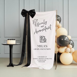 Könnte beinhalten: Ein Banner für eine Abschlussfeier mit dem Text "Offiziell ein Buchhalter" und "Mylas Abschlussfeier". Das Banner ist weiß mit schwarzem Text und einer schwarzen Schleife. Ein kleiner Kuchen steht auf einem schwarzen Tisch. Ballons in Schwarz, Weiß und Beige sind rechts.