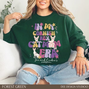 May include: Forest green sweatshirt with a graphic design that says "In My Cornish Rex Cat Mom Era" with a paw print and two cartoon cats. The text is in a retro style font with pink, purple, yellow, and blue colors.