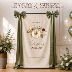 May include: A beige fabric sign hanging on a black metal stand, adorned with two large olive green satin bows. The sign features a printed illustration of a place setting with text reading Welcome to Thanksgiving Dinner 14th November 2026 New York.