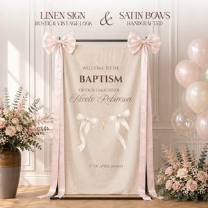Pode incluir: Uma placa retangular de linho bege pendurada num suporte de metal preto, com o texto "WELCOME TO THE BAPTISM OF OUR DAUGHTER NICOLE ROBINSON" e "1ST OF APRIL, MIAMI". Dois grandes laços de cetim rosa pálido estão presos aos cantos superiores.