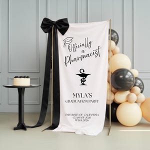May include: A graduation banner with the text "Officially a Pharmacist" and "Myla's Graduation Party." The banner is decorated with a black bow and black ribbons. A small cake and balloons are also visible.