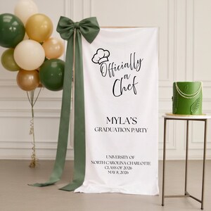 Könnte beinhalten: Eine Abschlussfeier-Szene mit einem weißen Banner mit der Aufschrift "Officially a Chef" und "Mylas Abschlussfeier". Ein grüner Kuchen mit Perlenakzenten steht auf einem kleinen Tisch. Ballons in Grün, Gold und Weiß sind zusammengebunden. Eine grüne Schleife und Bänder schmücken das Banner.