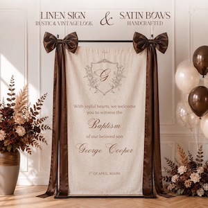 Puede incluir: Un letrero rectangular de lino beige colgado de un soporte de metal negro, con lazos de satén marrón oscuro en las esquinas superiores. El letrero muestra un escudo con la letra G, el texto "With joyful hearts, we welcome you to witness the Baptism of our beloved son George Cooper" y "1st of April Miami".