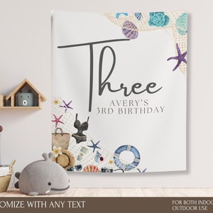 Może przedstawiać: Biały baner z napisem "Three" i "Avery's 3rd Birthday" w czarnym skrypcie. Baner jest ozdobiony akwarelowymi ilustracjami muszli, rozgwiazd, kostiumu kąpielowego i torby plażowej. Tekst na dole mówi "Customize with any text" i "For both indoor and outdoor use."