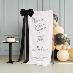 May include: A graduation party banner with the text "Officially a Software Engineer" and "Myla's Graduation Party." The banner is decorated with a black bow and black ribbons. A small cake sits on a black table. Balloons in black, white, and tan are on the right.