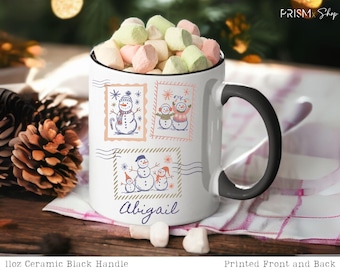 May include: White ceramic mug with a black handle, filled with marshmallows. The mug features postage stamp-style illustrations of snowmen and the name "Abigail". The mug is 11oz.