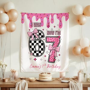 Op de afbeelding: Een witte verjaardagsbanner met roze glazuur, de tekst "I WAS 6, NOW I'M 7" en "Emma's 7th Birthday". De banner heeft een geblokte nummer 6 en een luipaardprint nummer 7, beide met roze strikken. Ballonnen en een taart zijn ook zichtbaar.