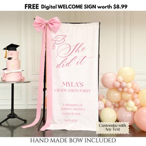 May include: A graduation party display with a pink banner reading "She did it" and "Myla's Graduation Party". A three-tiered pink cake with graduation caps is on a small table. Pink balloons and a handmade bow are also present.
