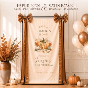 Peut inclure: Une bannière en tissu crème portant l'inscription 'OUR LITTLE PUMPKIN IS ONE!' et 'WELCOME TO Jackson's FIRST BIRTHDAY' avec une illustration de citrouille. La bannière est décorée de deux grands nœuds en satin brun rouille brillants.