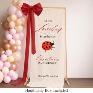 Puede incluir: Una pancarta de baby shower con el texto "A Little Lovebug is on the way!" y "Caroline's BABY SHOWER". La pancarta tiene una ilustración de mariquita roja y está decorada con un gran lazo rojo y un arco de globos en tonos rosa, melocotón y dorado.