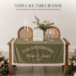 Op de afbeelding: Olijfgroene satijnen kanten tafelloper met witte kanten rand, gepersonaliseerd met "The Andersons" en "Olivia & James, EST. 2026". De loper ligt op een houten tafel, versierd met kaarsen, bloemen en wijnglazen. De tekst "SATIN LACE TABLE RUNNER" staat bovenaan.