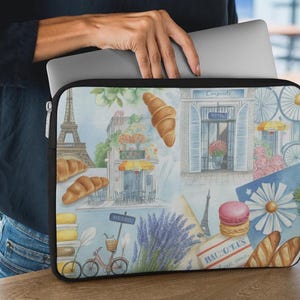May include: A rectangular laptop sleeve featuring a watercolor print of Parisian scenes, including the Eiffel Tower, croissants, a bicycle, lavender, macarons, and baguettes. The sleeve has a black border, a silver zipper, and a soft interior lining.