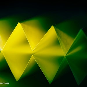 May include: Abstract image of a folded paper design in a zigzag pattern. The paper is yellow, fading to green in the shadows, against a dark green background. The central design is in sharp focus, creating a striking visual.